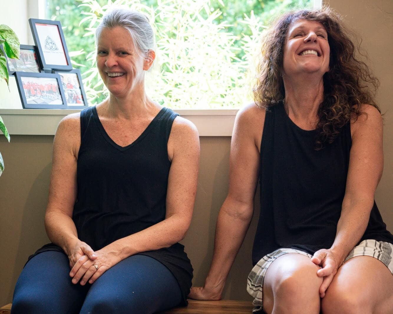 Two women on the studio bench, laughing together in the window light.
